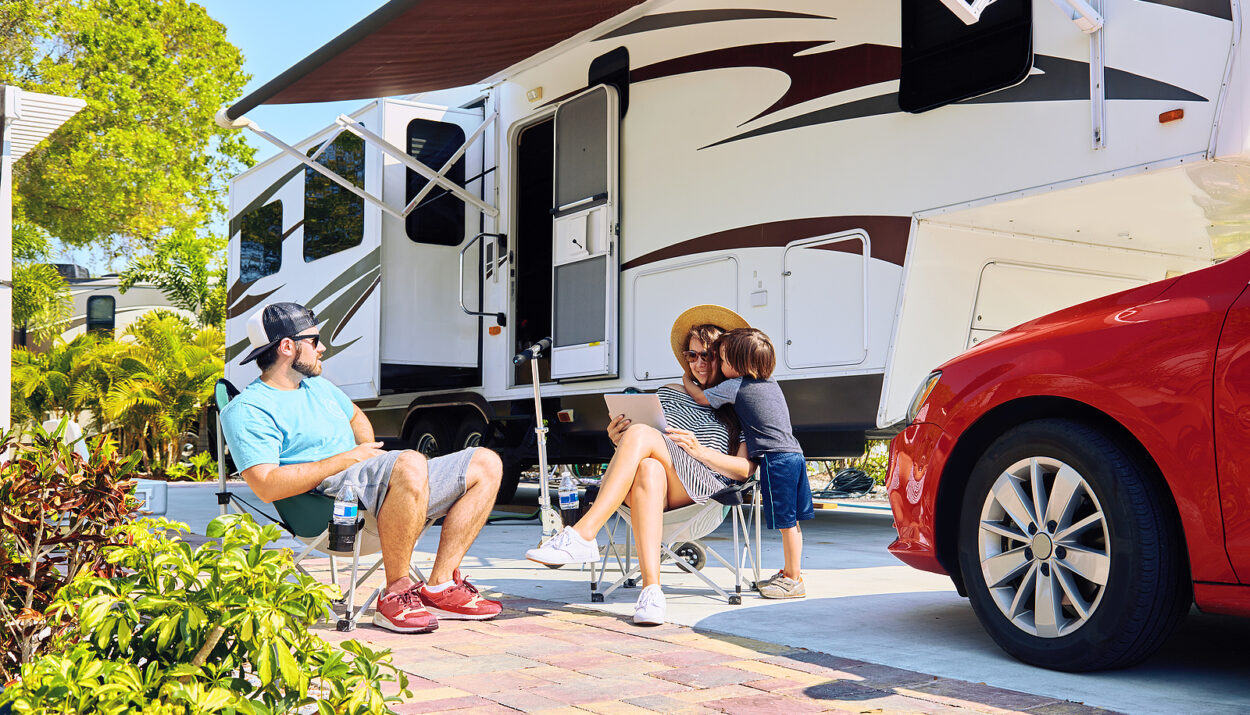 Mother father and son sitting near camping trailer smiling. Woman men kid relaxing on chairs near car and palms. Family spending time together on vacation near sea or ocean in modern rv park