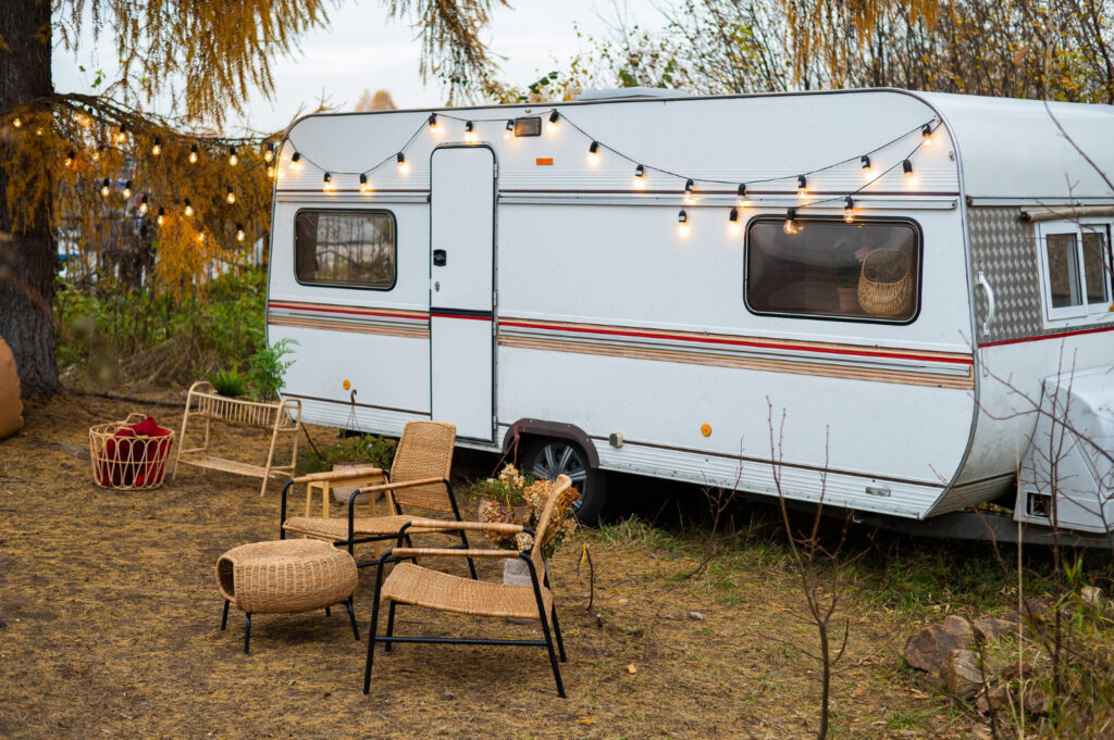 A white with red and orange stripes camper outside in nature with a few chairs and lights from the ceiling 