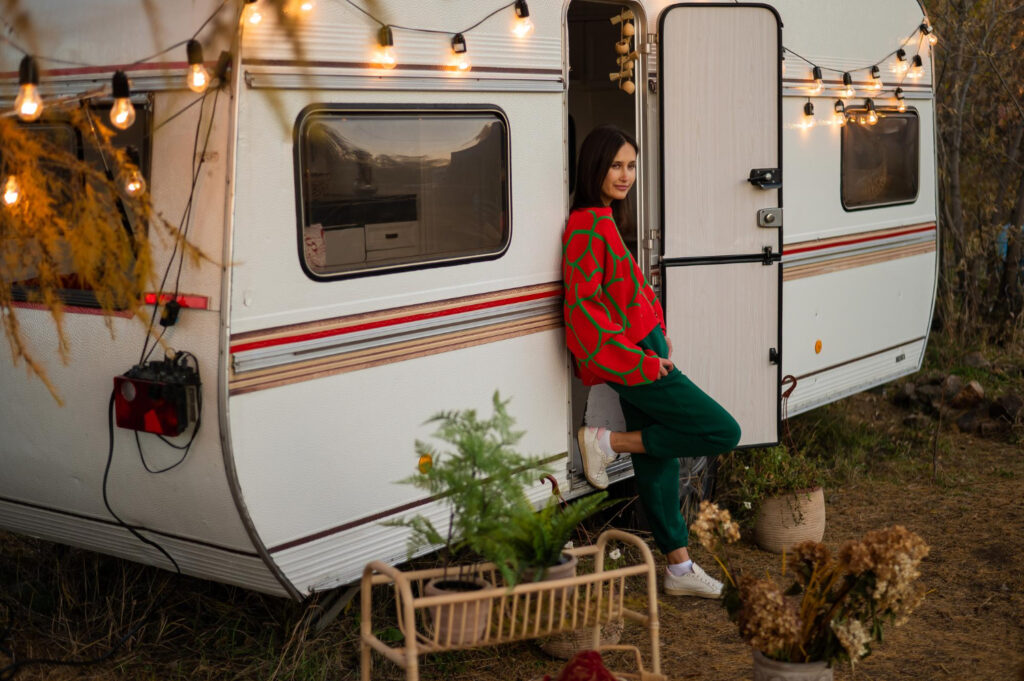 Woman in a red and green sueter enjoying a day in nature next to a white with red and blue stripes camper 