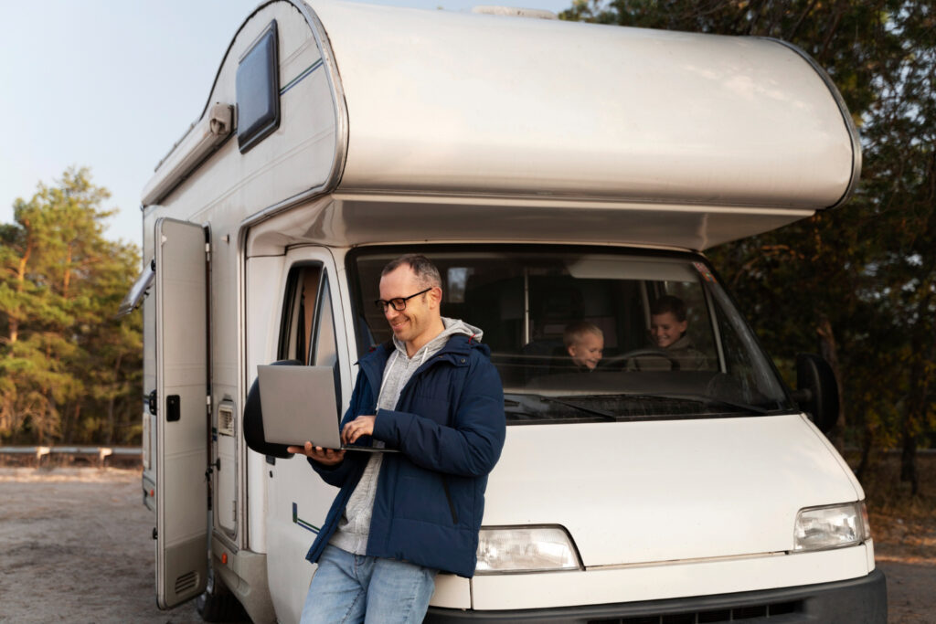 Man in a blue jacket and a grey sueter holding a laptop in front of a white camper in the forest with two childrens in the front seat