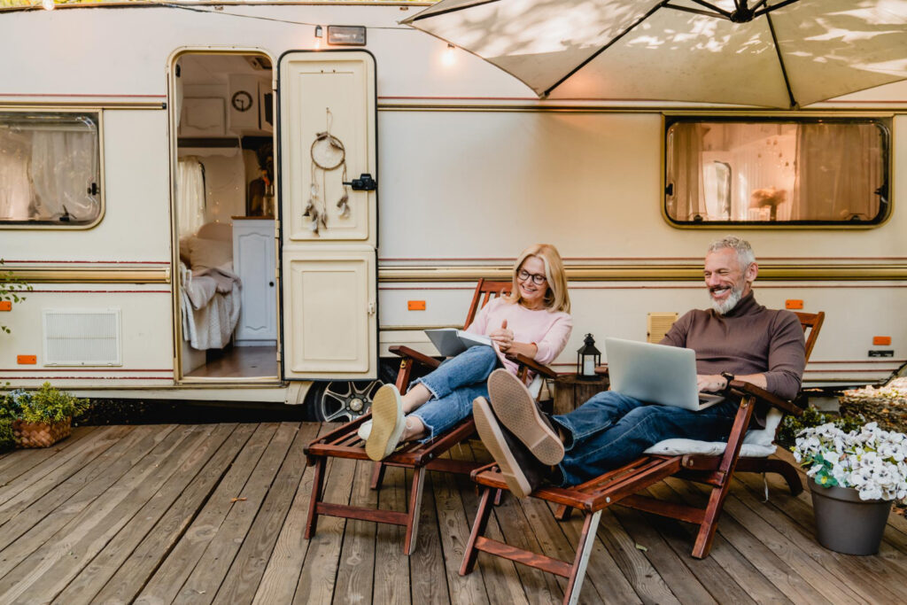 A couple of campground owners in a brown and beige sueter outside of a camper with a book and a laptop managing their business