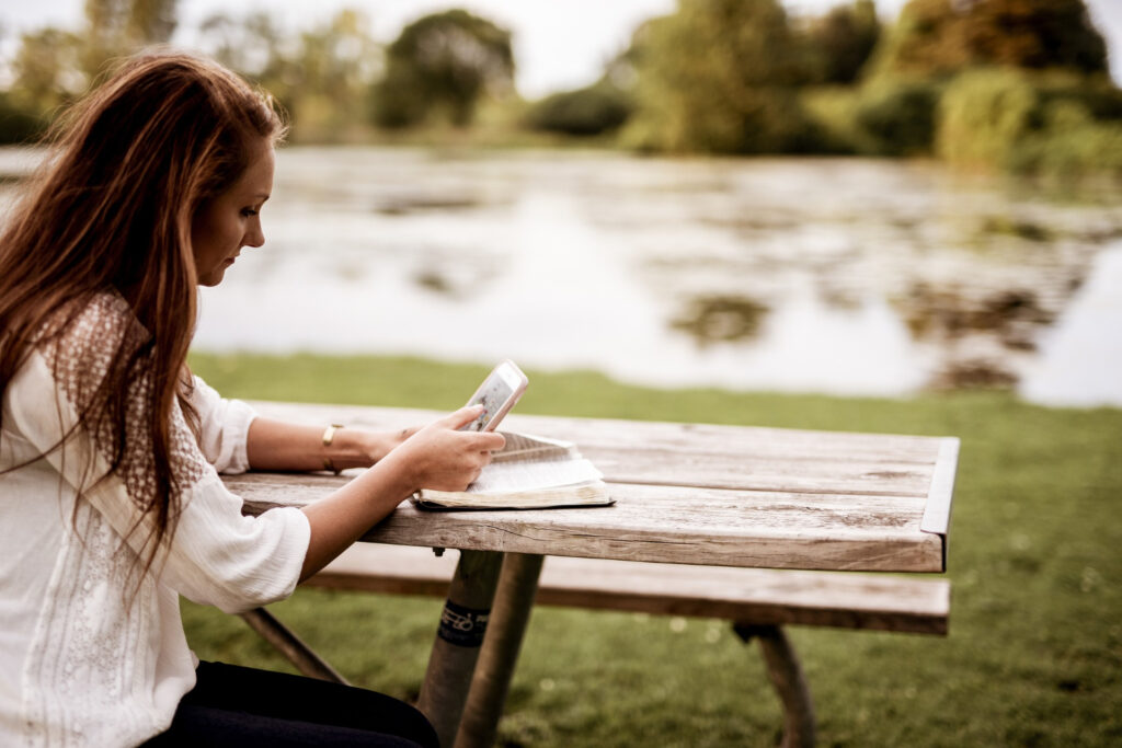 A woman in a white shirt sitting in a wooden chair while checking her phone to see her campground availability