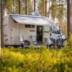 A white and grey camper van with two chairs outside in a low point of view at the forest