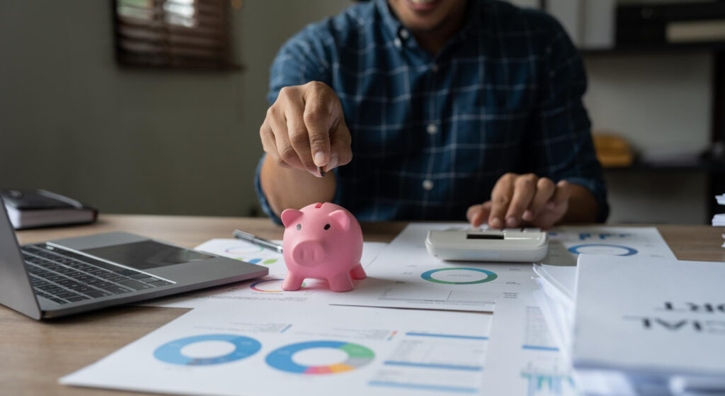 A man with a blue button shirt saving money inside of a pig with a laptop and some reporting papers in his table