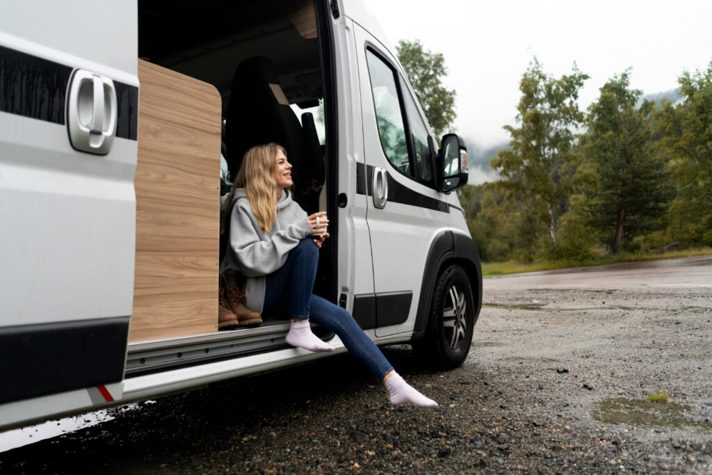 A woman in a grey sueter sitting in her white with black stripes camper van enjoying a coup of coffee in nature