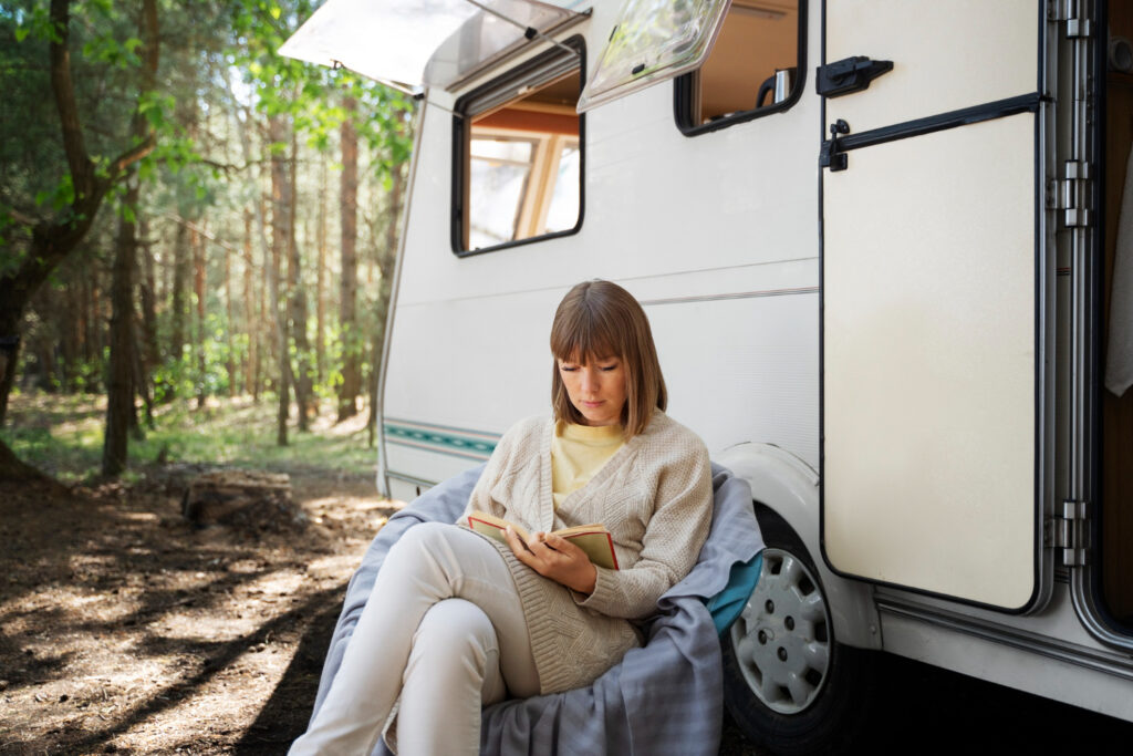 a woman in a beige cardigan and yellow shirt reading in nature outside of a white camper 