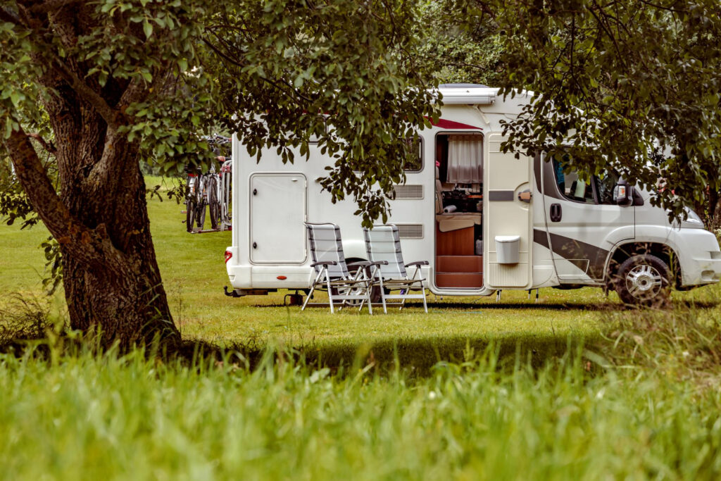 a white and grey camper in the forest with two chairs outside of a RV park 