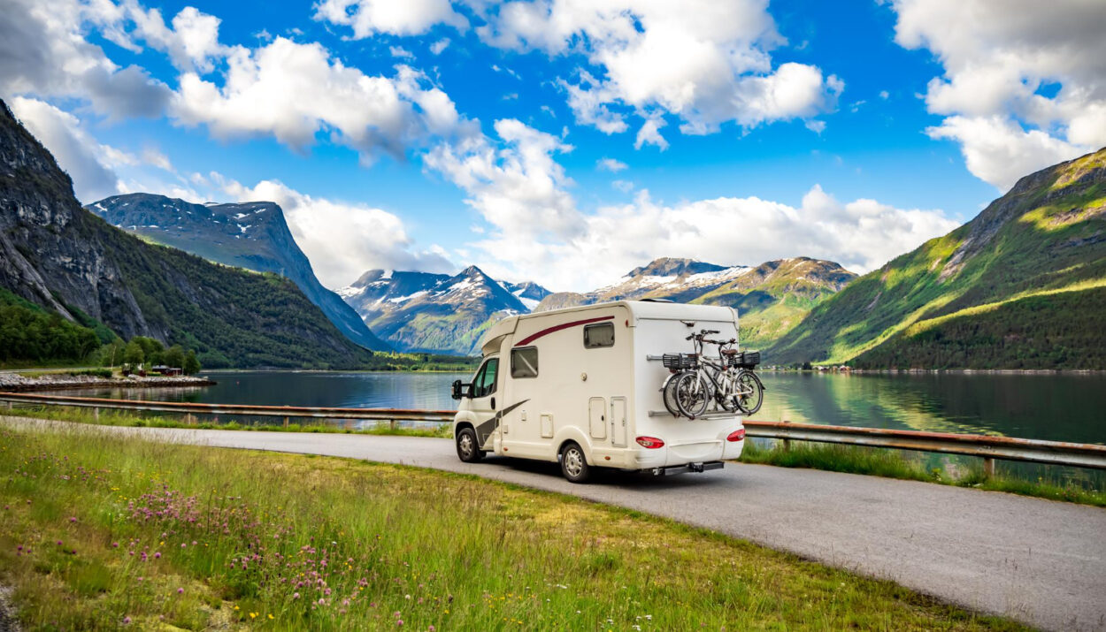 A white camper van with two bikes attached behind it going on the road with mountain on their side and a blue sky