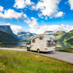 A white camper van with two bikes attached behind it going on the road with mountain on their side and a blue sky