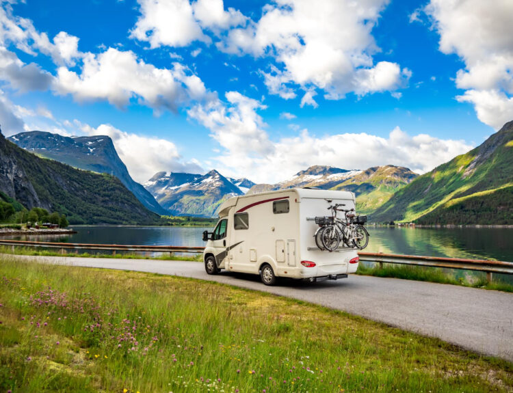 A white camper van with two bikes attached behind it going on the road with mountain on their side and a blue sky