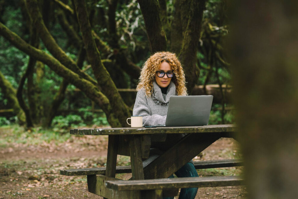 Woman with grey sueter in the forest using her laptop to check her software reservation system