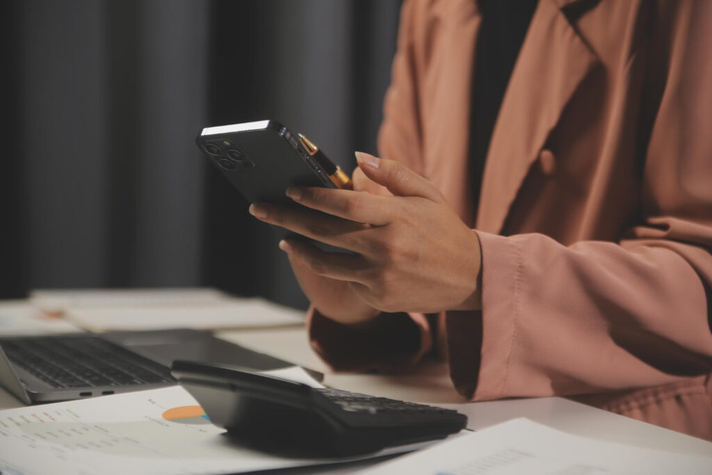 A woman with a salmon coat using her phone with a laptop and a calculator in her table seeing the payment options of a campground