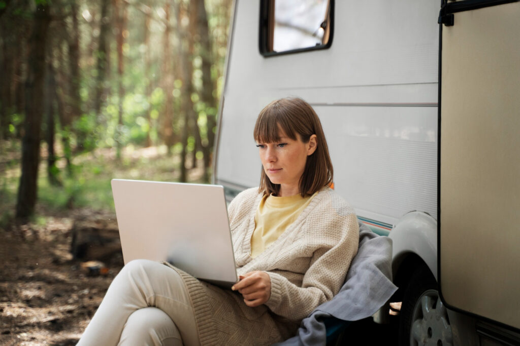 woman sitting outside of a camper van using a grey laptop booking a new campground spot using campground booking software