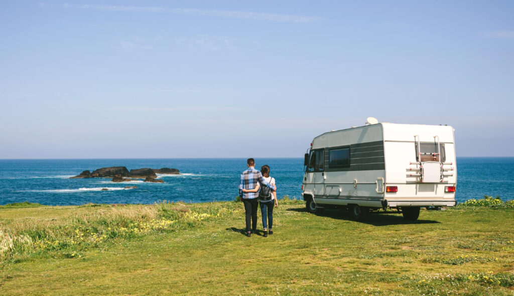 a couple with a white and grey camper van in front of the ocean 