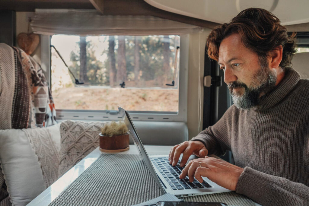 Man with a grey sueter sitting inside his camper van using a laptop to make a reservation using a campground software 