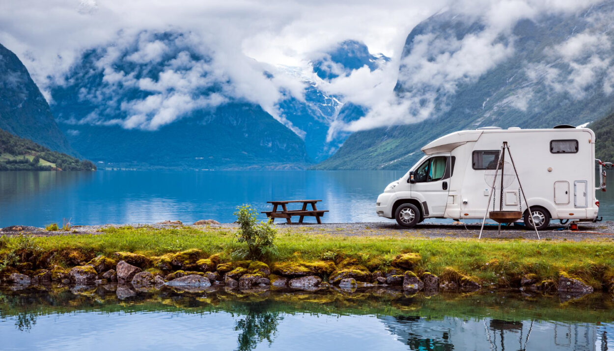 A white camper van in front of a lake with their reflection in the water with a few mountains