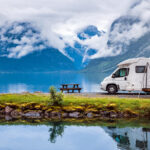 A white camper van in front of a lake with their reflection in the water with a few mountains