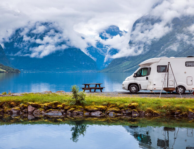 A white camper van in front of a lake with their reflection in the water with a few mountains