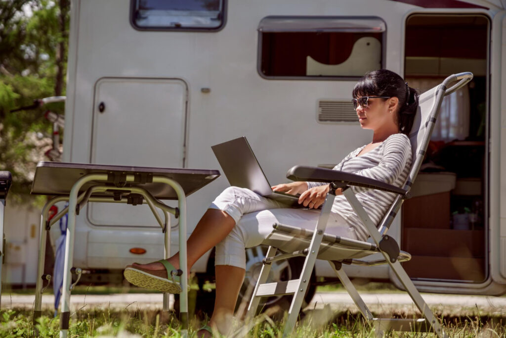 a woman with a white sueter with horizontal grey lines using her laptop outside of her camper van creating a marketing campaign for her RV park using campground management software