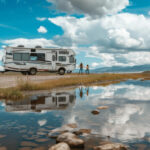 a camper van staying in front of a lake with its reflection and two poeple outside of it enjoying the view